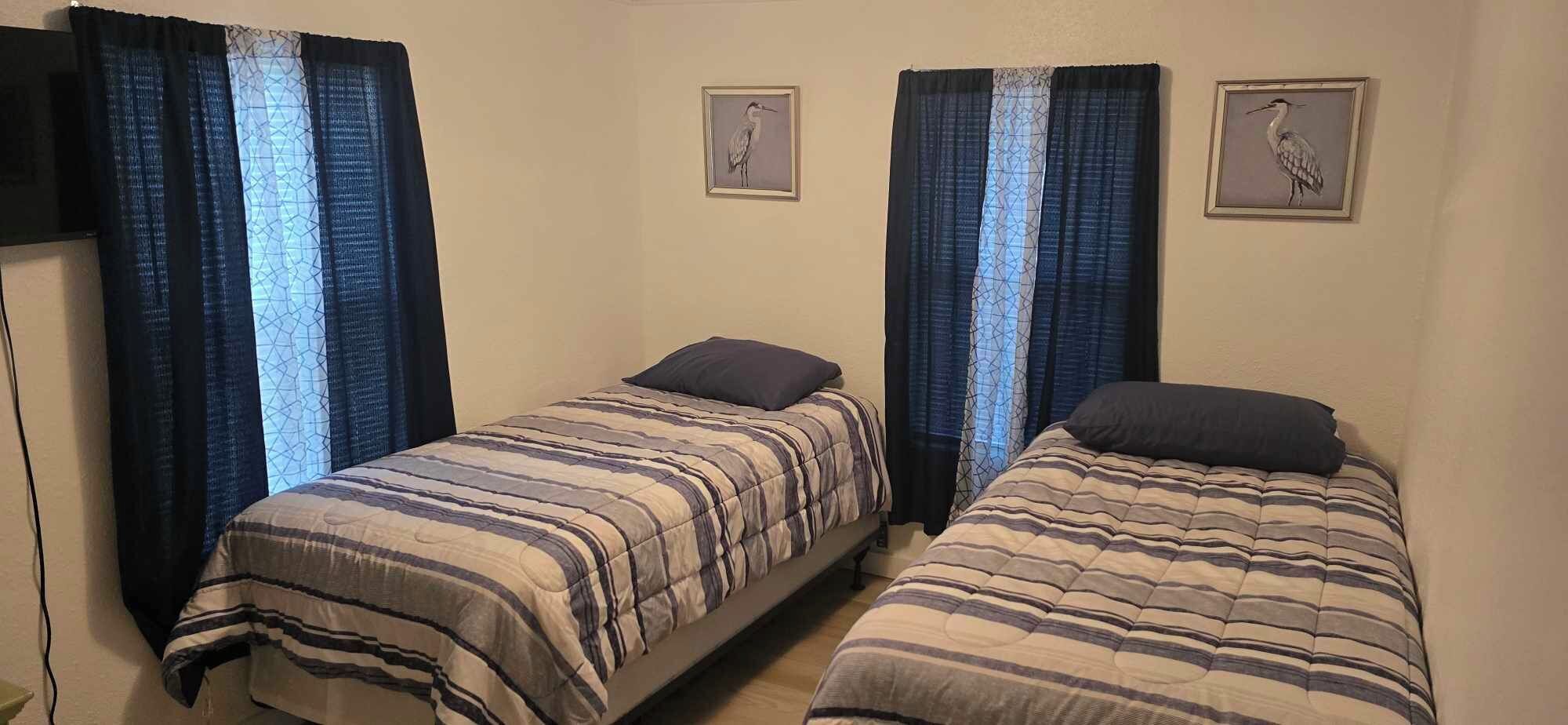 A room with two single beds, each with a striped bedspread and a dark pillow. There are two framed pictures on the wall, one above each bed. The room has a simple, functional design with plain walls and a window with blue curtains.
