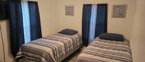 A room with two single beds, each with a striped bedspread and a dark pillow. There are two framed pictures on the wall, one above each bed. The room has a simple, functional design with plain walls and a window with blue curtains.