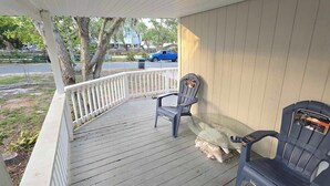 A covered porch with two chairs and a table, a white railing, and a view of a street with a blue truck. - Clambassador Cottage (Cedar Key)