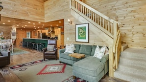 A spacious living area with a wooden staircase, a green sofa, and a kitchen in the background. - County Highway - Trail Access, Lake Access, Sleeps 23 (Minong)