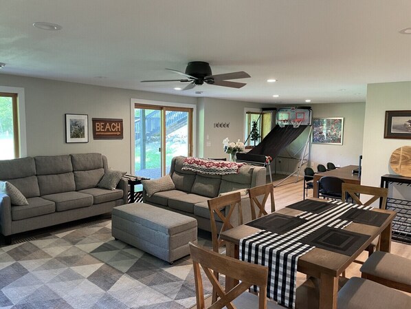 A living room with a sofa, ottoman, dining table, and chairs. There is a ceiling fan, a basketball hoop, and a staircase leading to another level.