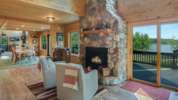 A cozy cabin interior with a stone fireplace, wooden furniture, and a large window overlooking a lake. - County Highway - Trail Access, Lake Access, Sleeps 23 (Minong)