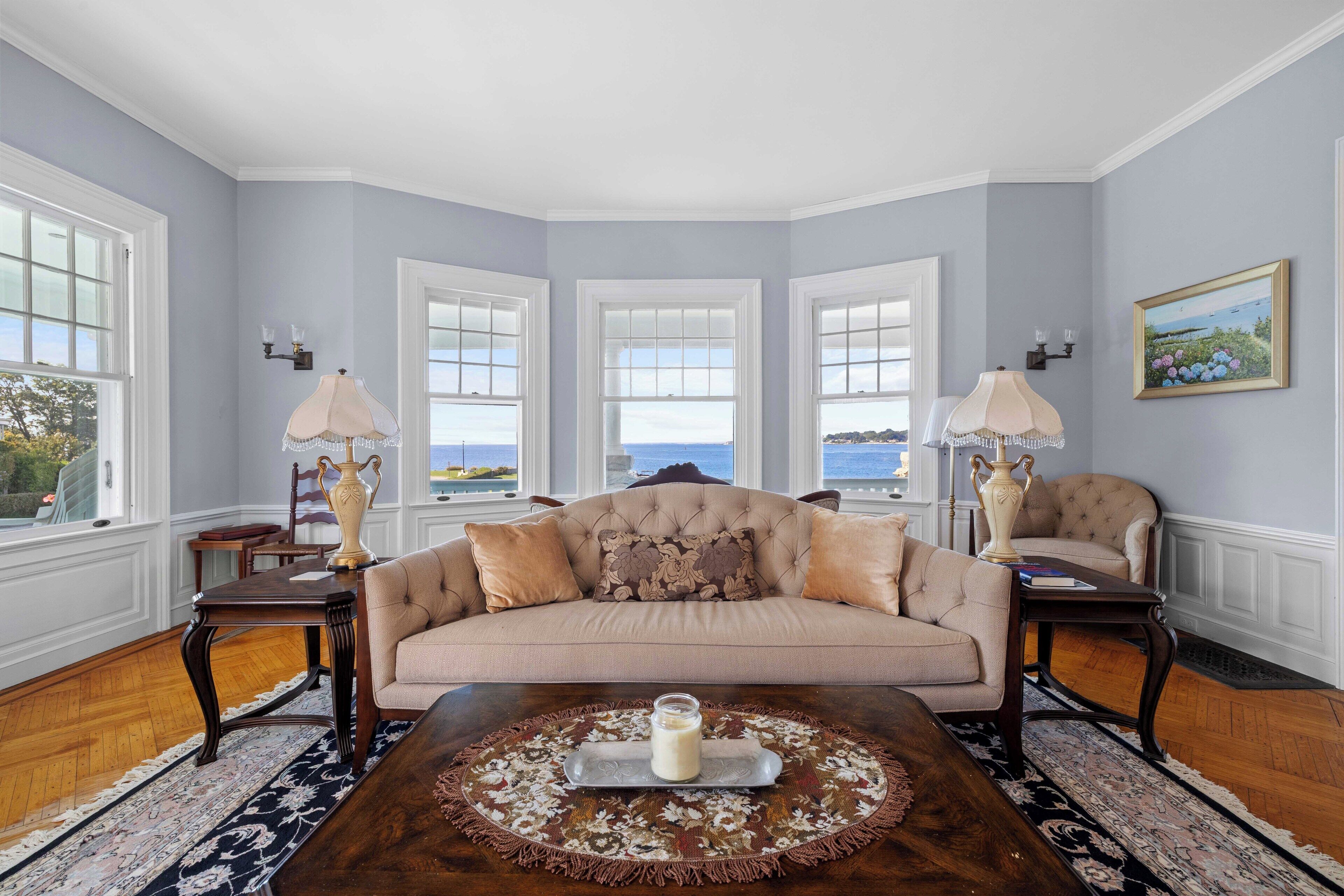 Un salon avec un canapé beige, une table basse agrémentée d’un tapis décoratif et une vue sur la mer grâce à de grandes fenêtres.