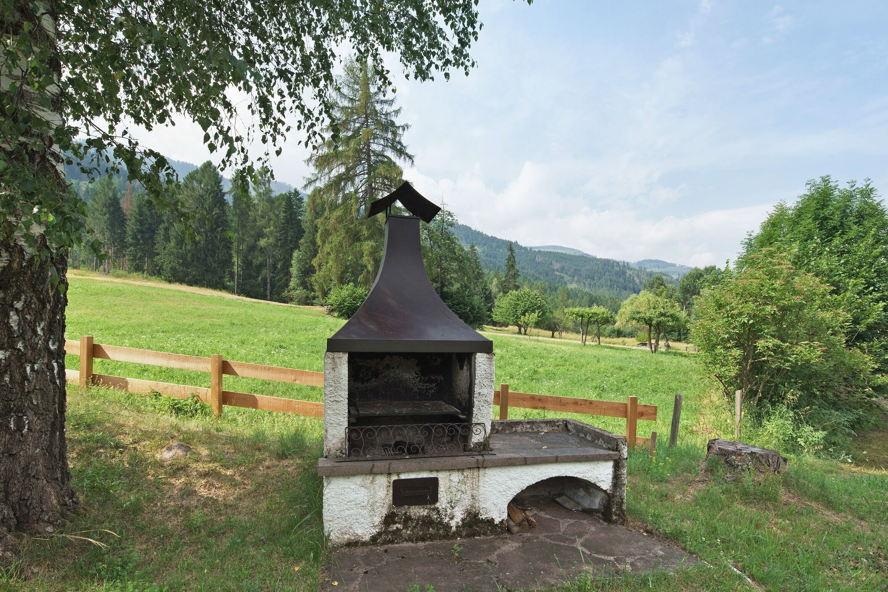 Ein rustikaler Grill im Freien mit Schornstein, umgeben von einem Holenzaun und viel Grün.