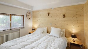 Une chambre à coucher avec une tête de lit en bois, deux tables de chevet avec des lampes, un radiateur et une fenêtre ornée de rideaux en dentelle.