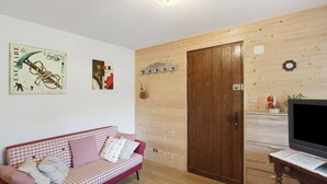 Un salon avec un canapé à carreaux, des murs en bois, une porte en bois, un téléviseur posé sur un meuble en bois et une horloge murale.