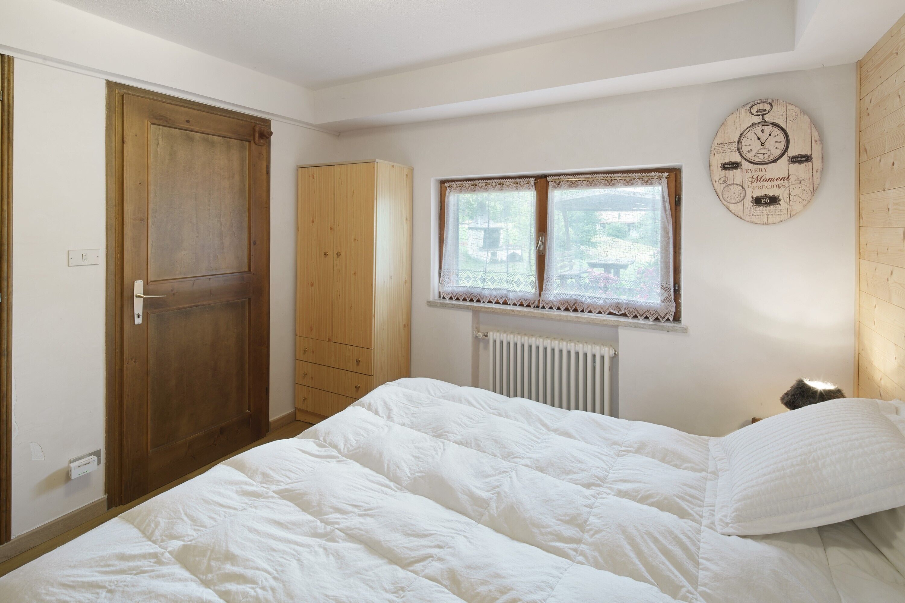 A bedroom with a bed, a wooden wardrobe, a window with lace curtains, and a wall clock.
