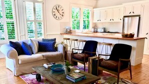 A cozy living room with a sofa, coffee table, and chairs, a kitchen in the background, and large windows with blinds.