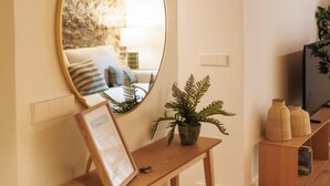 Een woonkamer met een ronde spiegel, een houten console, een lijst met een afbeelding, een potplant varens, een televisie en een boekenplank.