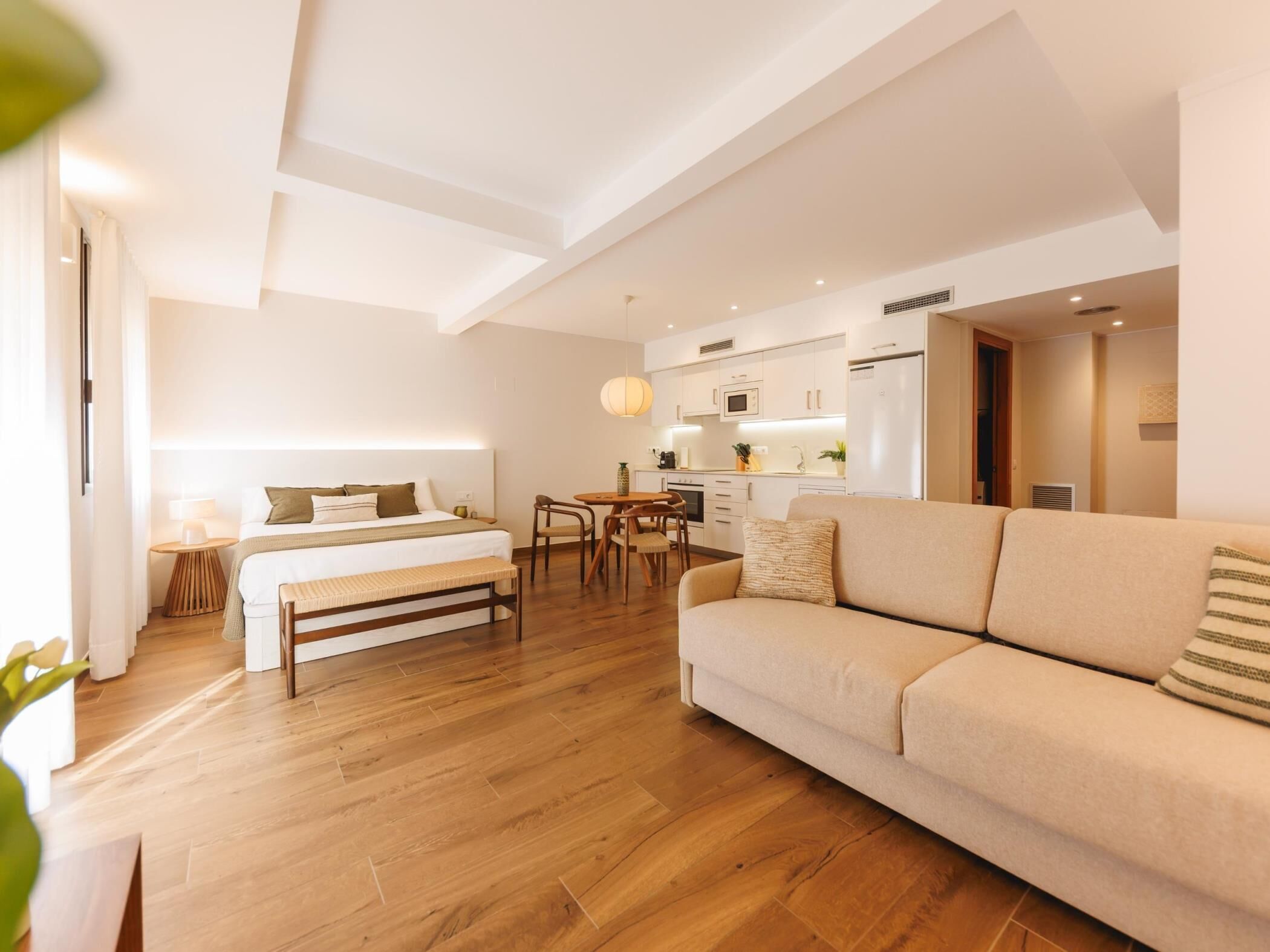 A modern hotel room with a large bed, a sofa, a dining area, and a kitchenette.
