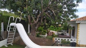 Una piscina amb un tobogan blanc, envoltada per un pati tancat amb arbres i un jardí petit.