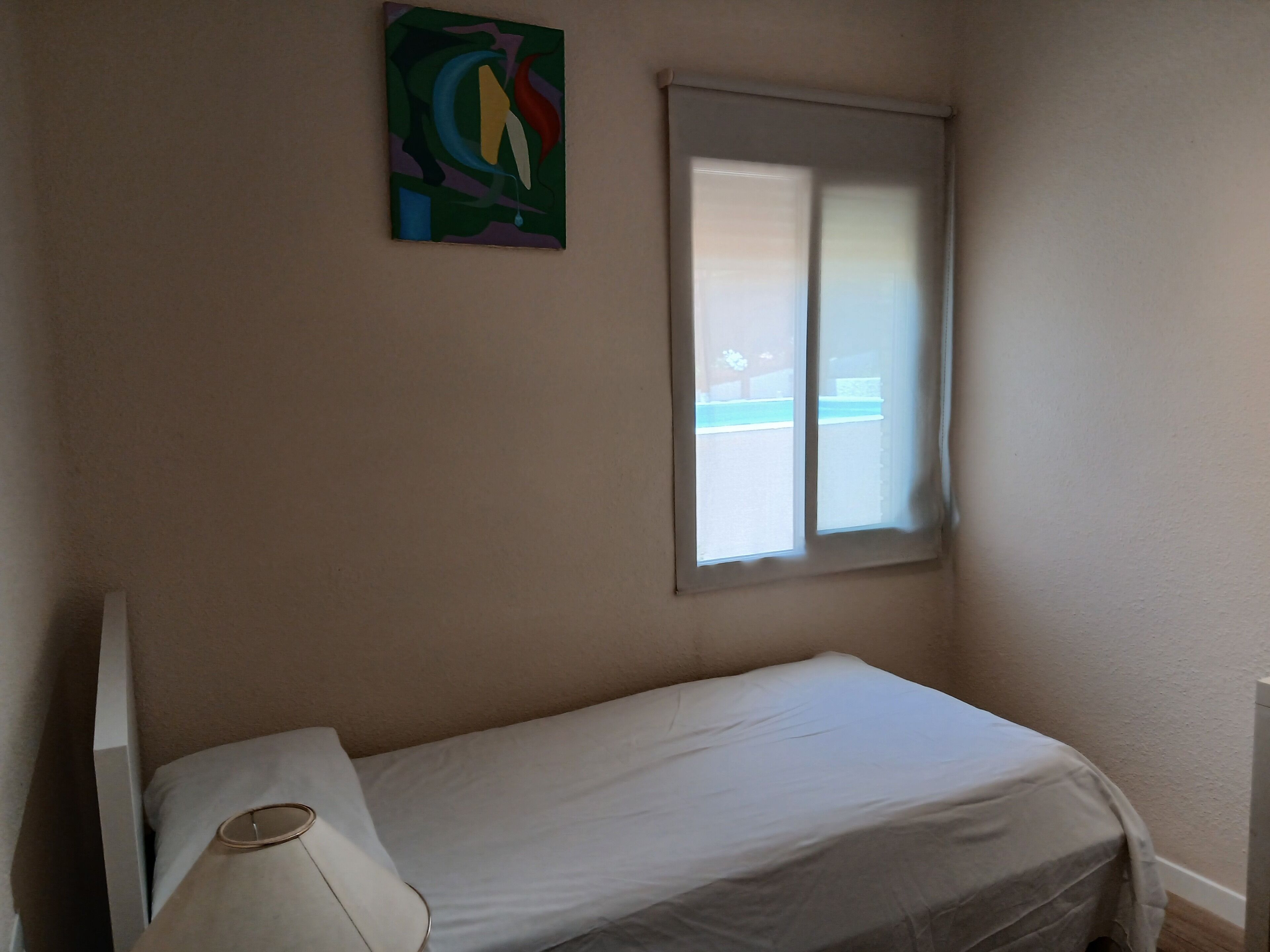 A small, single-bed room with a window, a wall-mounted painting, and a bedside lamp.