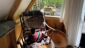 A cozy attic bedroom with a wooden ceiling, a window with curtains, a wooden chair, a small table, and a view of the outdoors.