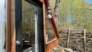 A wooden cabin with a black door, a small porch with a wooden bench, and a view of a forested area. - Hillside: Cozy offgrid cabin near Cooperstown, NY (Cherry Valley)