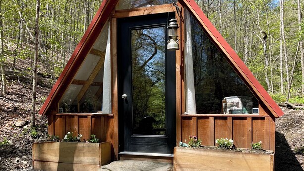 A small, triangular wooden structure with a black door, wooden benches, and a potted plant in the foreground. - Hillside: Cozy offgrid cabin near Cooperstown, NY (Cherry Valley)