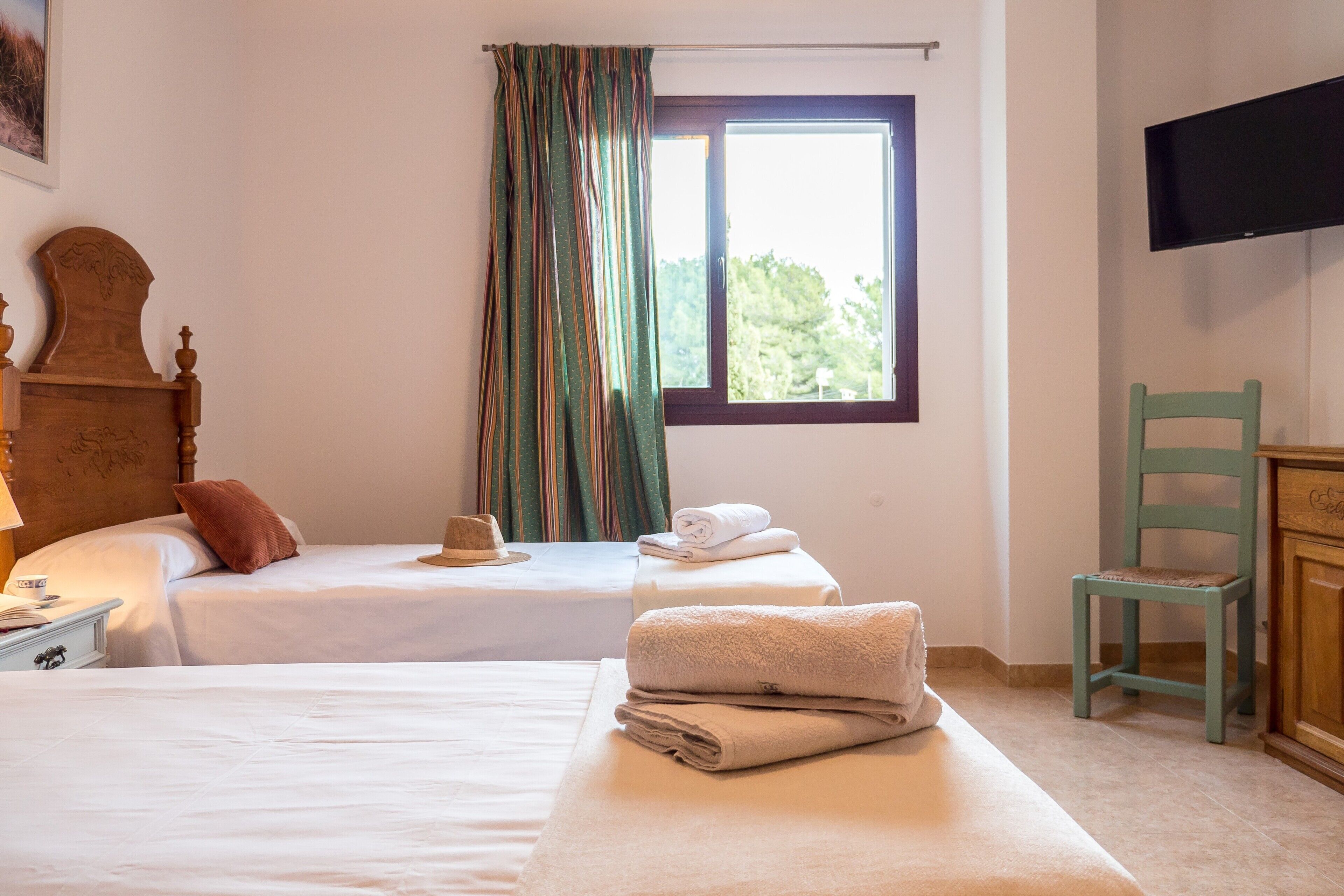 Ein Hotelzimmer mit zwei Betten, einem fensterbelaubten Fenster, einem an der Wand montierten Fernseher und einem Holzstuhl.