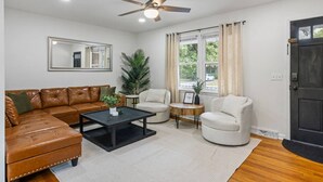 A living room with a brown leather sofa, a black coffee table, a white armchair, and a small side table. There is a ceiling fan, a window with curtains, and a door.