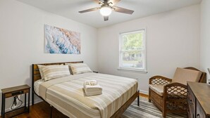 A bedroom with a bed, a chair, a desk, a ceiling fan, a window, and a painting on the wall.