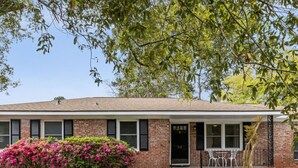 A two-story brick house with a front porch, a small garden with pink flowers, and a tree in the background.