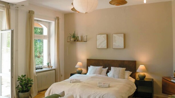 Une chambre avec un grand lit, deux tables de chevet, une fenêtre avec des rideaux, un radiateur et des étagères murales ornées de plantes.