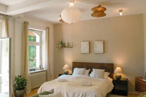 Een slaapkamer met een groot bed, twee nachtkastjes, een raam met gordijnen, een radiator en wandplanken met planten.