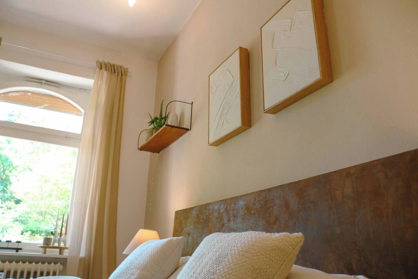 Una camera da letto con un letto, due quadri appesi al muro, una pianta sul comodino e una finestra con tende.