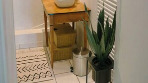 Un couloir étroit avec un sol carrelé de carreaux blancs, une table en bois surmontée d’un lavabo blanc et deux paniers en osier. On y trouve deux plantes en pot, une posée sur le sol et l’autre accrochée au mur.