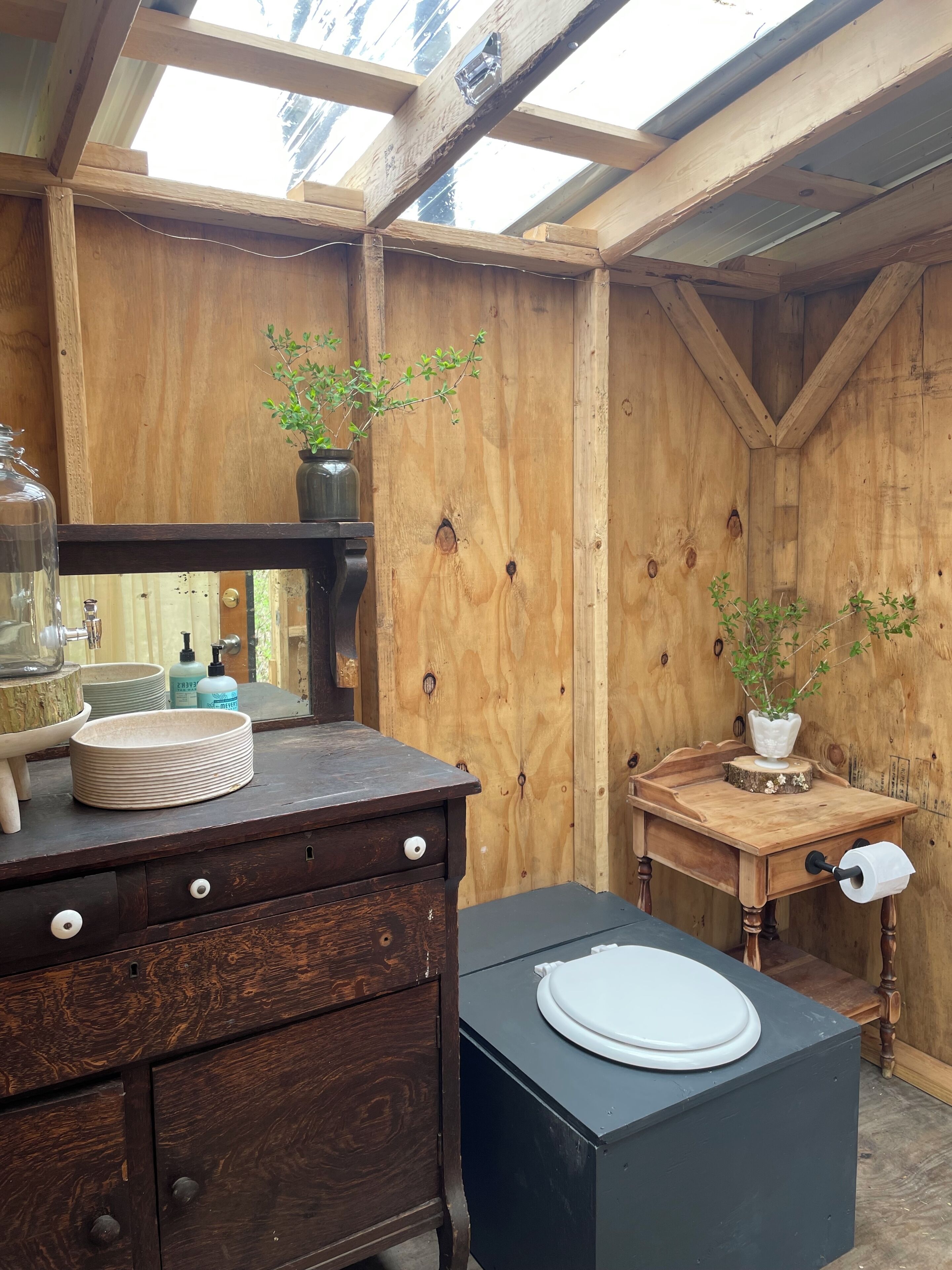 Una habitació de fusta amb un finestral i un escriptori de fusta amb un dispensador de paper higiènic, i dues plantes en testos.