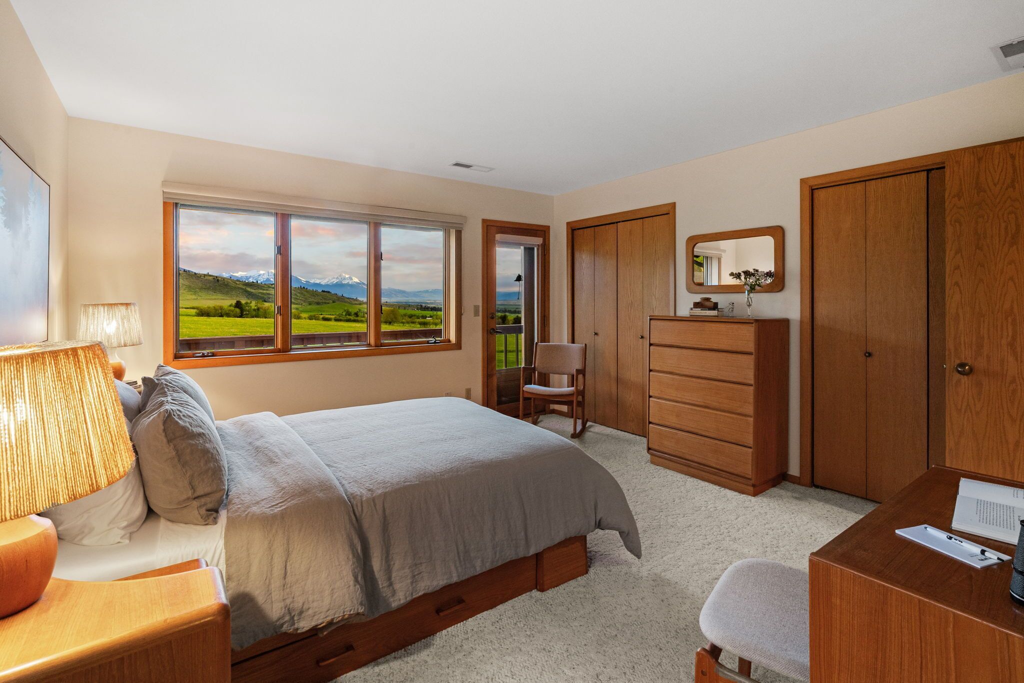 A bedroom with a large bed, a desk, a chair, a dresser, and a window with a scenic view.
