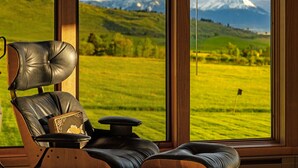 A leather chair with a cushion and a small table, positioned by a large window with a view of a green field and mountains. - Spacious home with mountain and valley views, seconds to Deep Creek trailhead (Livingston)