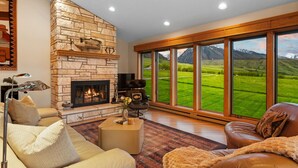 A spacious living room with a stone fireplace, leather sofas, and large windows offering a view of a green landscape. - Spacious home with mountain and valley views, seconds to Deep Creek trailhead (Livingston)