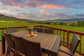 A wooden deck with a table set for dining, surrounded by wicker chairs, overlooking a green landscape with distant mountains. - Spacious home with mountain and valley views, seconds to Deep Creek trailhead (Livingston)