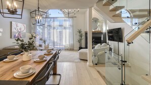 Uma área de jantar moderna com uma mesa de madeira posta para quatro pessoas, uma escada de vidro e uma janela grande com cortinas leves.