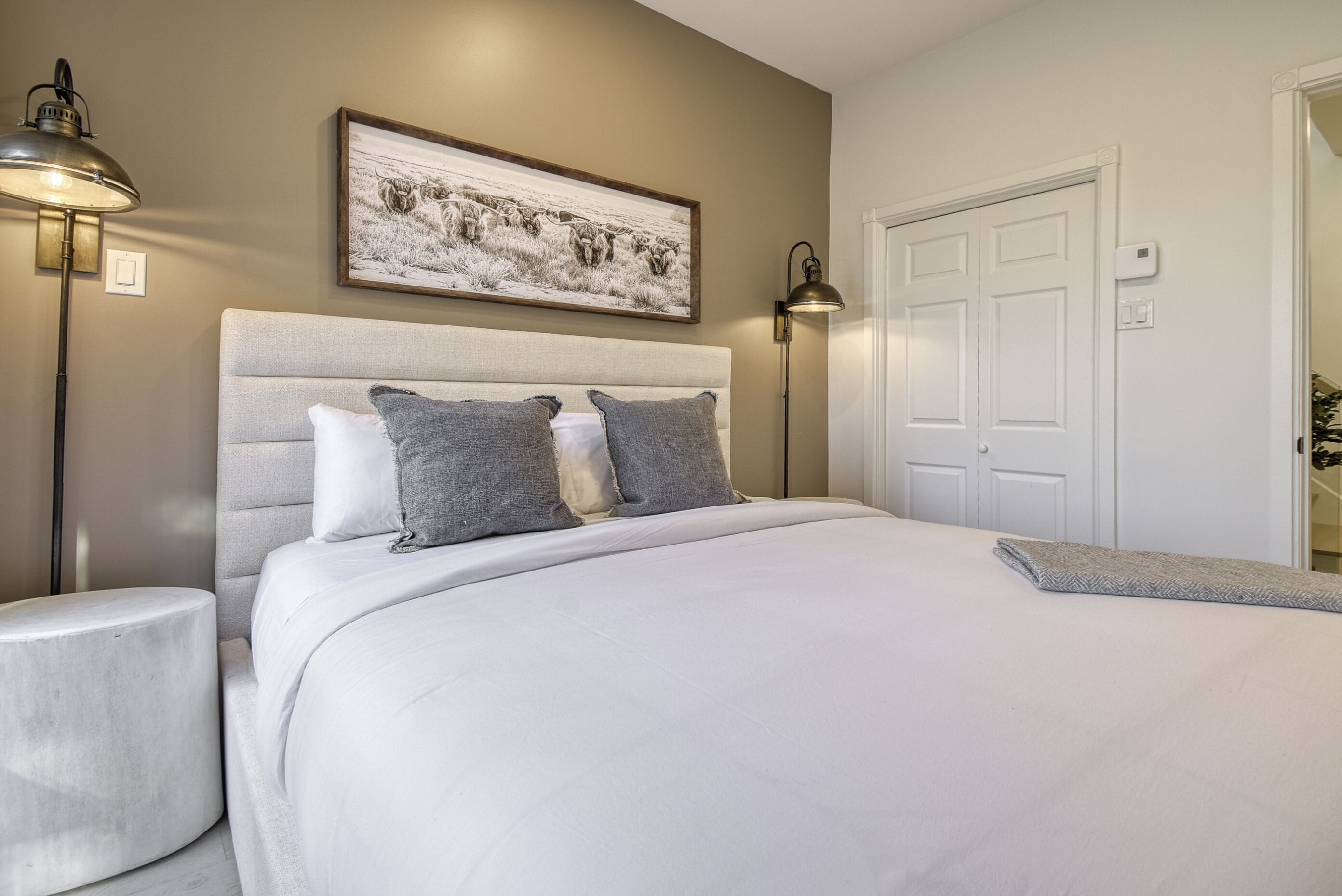 A bedroom with a large bed, a headboard, two bedside lamps, a framed picture on the wall, and a white door.