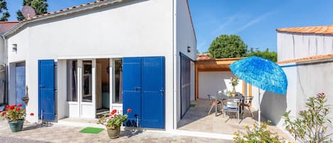 A white building with blue shutters, a patio with a table and chairs, and potted plants.