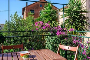 A balcony with a wooden table and chairs, surrounded by lush greenery and purple flowers.