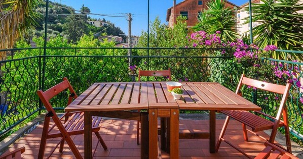 A wooden table and chairs on a balcony with a green metal railing, surrounded by lush greenery and flowers.