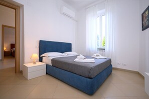 A bedroom with a blue bed, white pillows, a grey blanket, a bedside lamp, and a window with curtains.