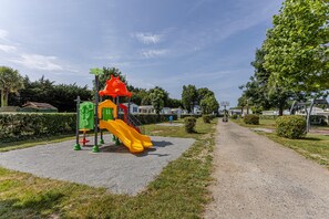 A playground with a slide and climbing structures, surrounded by a gravel path and greenery.