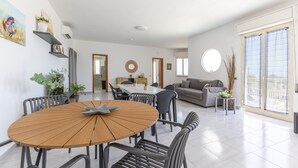 A modern living room with a round wooden dining table, grey chairs, a grey sofa, and a large window with blinds. - Villa 'Primavera' with sea view, private terrace and air conditioning (Marina di Modica)