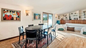 Un salon moderne avec un coin repas, comprenant une table ronde, des chaises noires et un tapis à motifs. Deux œuvres d’art encadrées ornent les murs, et une bibliothèque contient divers objets.
