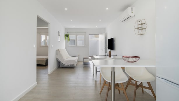 Une cuisine moderne avec des meubles blancs, un coin repas avec une table et des chaises blanches, et un salon avec un canapé blanc et une plante.
