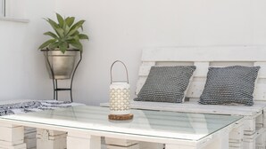 Une table à plateau en verre, agrémentée d’une lanterne et d’une plante en pot, placée à côté d’un banc en bois blanc orné de coussins à motifs.