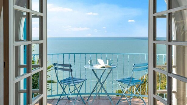 Un balcone aperto con vista sul mare, due sedie blu e un tavolino.