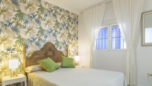 Une chambre à coucher comprenant un lit, une table de chevet, une lampe, une fenêtre avec des rideaux et un mur orné d’un motif de feuilles tropicales.