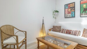 Un salon comprenant un fauteuil en osier, une table basse en verre, un canapé blanc agrémenté de coussins à motifs et des œuvres d’art murales.