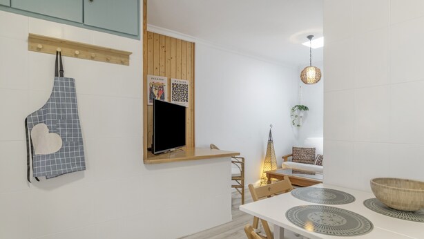 Une cuisine moderne avec un plan de travail en bois, un téléviseur fixé au-dessus, et un coin repas comprenant une table et des chaises blanches.