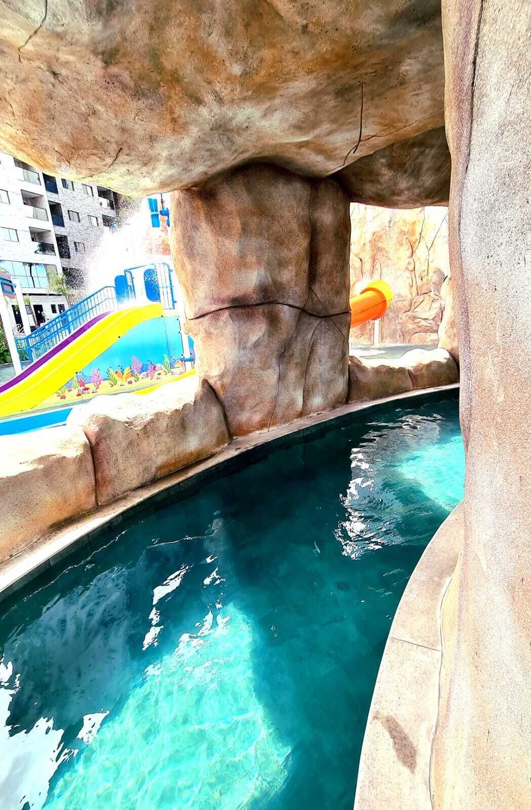 Un espace piscine agrémenté d’une grande formation rocheuse au-dessus, d’un toboggan coloré et d’une piscine à l’eau d’un bleu limpide.