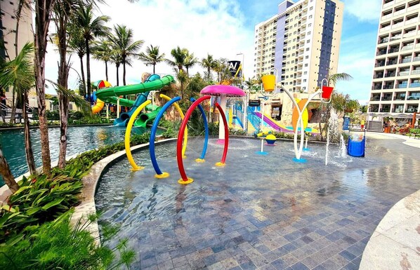 Parque acuático con toboganes y fuentes de agua, rodeado de palmeras y una piscina.