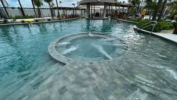 Una piscina con un área circular de hidromasaje, rodeada de palmeras y una zona de estar techada.